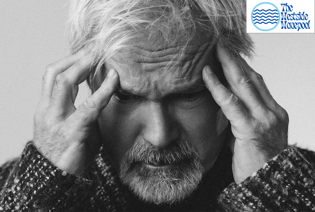 A close-up, black-and-white image of a man with gray hair and a beard, holding his head with both hands, conveying a sense of stress or discomfort.
